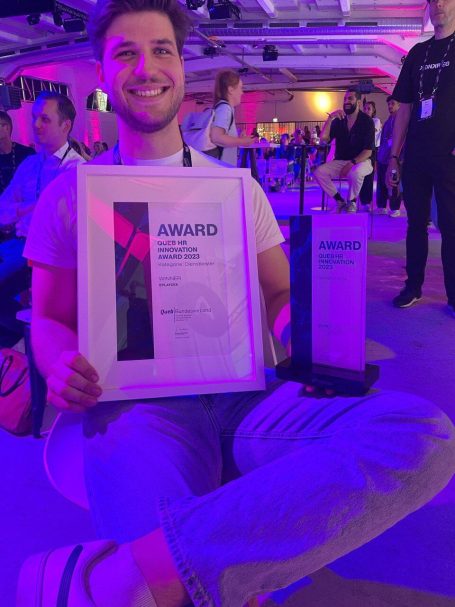 Junger Mann lächelt mit Preis und Urkunde in einer bunten Event-Umgebung.