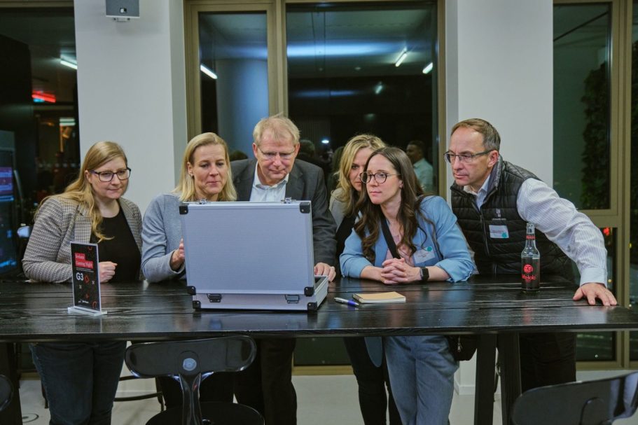 Gruppe von sechs Personen, die auf einen Rätselkoffer in einem modernen Raum schauen.